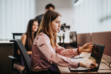 A professional woman in a pink blouse focuses intently on her laptop at a vibrant office workspace, colleagues in the background.