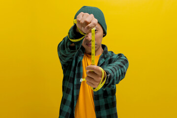 An Asian man, dressed in a beanie hat and casual clothes, measures his height using a yellow measuring tape while standing against a yellow background