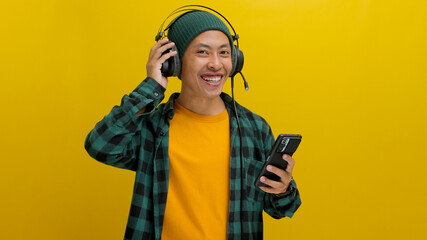 Asian man in a beanie and casual clothes listens to music on his headphones, phone held in hand. Isolated on a yellow background.