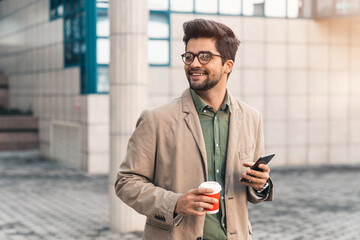 Young businessman with eyeglasses looking away while holding mobile phone and takeaway coffee...