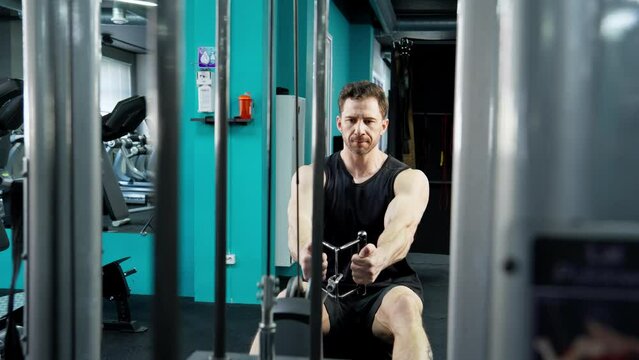 Man in tank top lifting blocks on rowing machine. Athletic man exercises back muscles on seated rowing machine in modern gym. Focused man working out indoors. Sport concept. 4K, UHD