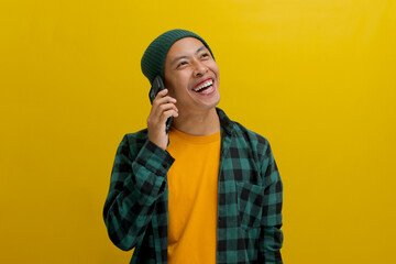 Cheerful young Asian man, dressed in a beanie hat and casual shirt, engages in a phone conversation, laughing and thoroughly enjoying a pleasant conversation while standing against a yellow background
