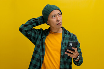 Pensive Asian man, dressed in a beanie hat and casual shirt, holds a mobile phone, appearing...