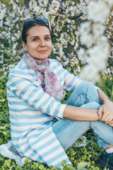 Young woman in striped cardigan and scarf sitting and posing on the background of apple flowers bloom
