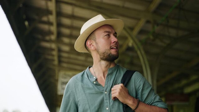 Asian traveler explores Haputale railway station with camera, backpack. Man in hat smiles, looks around historic site, embarks on solo journey. Authentic, rustic vibe of Sri Lanka travel adventure.