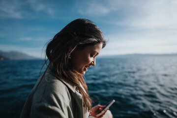 Naklejka premium Happy girl with a joyful expression using her phone by the seaside on a sunny day, evoking feelings of happiness and contentment.