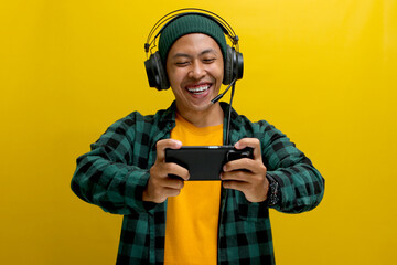 Excited young Asian man, wearing headphones, a beanie hat and casual shirt, engages in playing an...