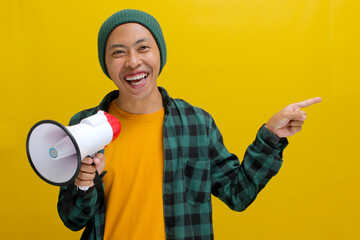 Happy Asian man in a beanie hat and casual shirt talks into a megaphone and points at empty copy...