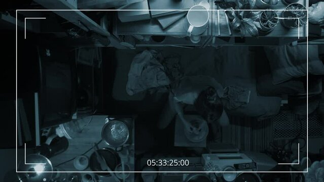 Full top surveillance camera footage of anonymous poor woman sitting on bunk bed in cramped shoebox apartment in evening, washing clothes by hand in plastic tub, water boiling on stove