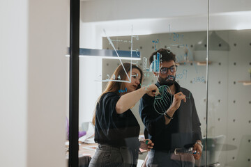 Two business professionals engaged in discussing and problem-solving while writing on a glass wall with sticky notes and markers in an office.