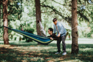 Two young men share a joyful moment as one swings in a hammock while the other assists, in a lush, sunlit park.