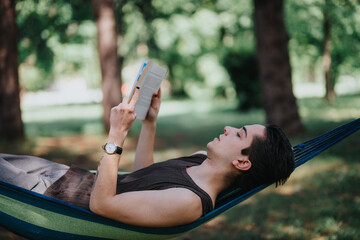 A young adult male relaxes with a book in a hammock, set against a lush green park background on a...