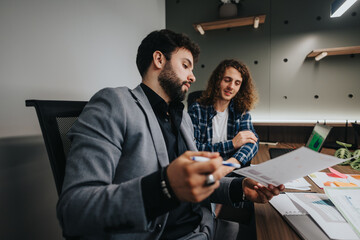 In a contemporary office setting, two male colleagues, one in a suit and another in casual attire, engage in a productive discussion over documents and diagrams.