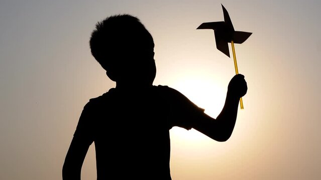 A boy stands under the hot sun and plays with a pinwheel fan. Silhouette view of an Asian child standing and spinning a handmade paper fan.