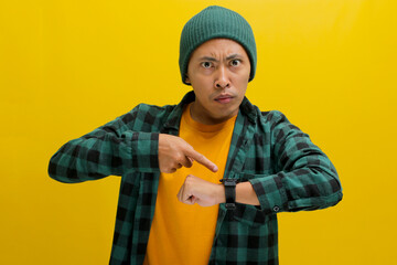 Frowning Asian man in a beanie and casual shirt impatiently taps his wristwatch, glancing at the...