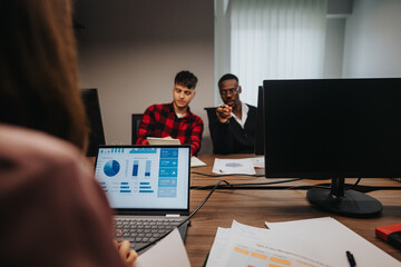 A diverse team of young businesspeople engaged in analyzing statistical data on a computer screen in a well-lit office environment.