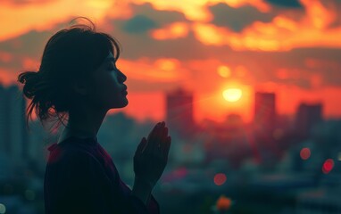 A woman is praying in front of a sunset. The sky is filled with clouds and the sun is setting. The woman is standing in front of a city skyline