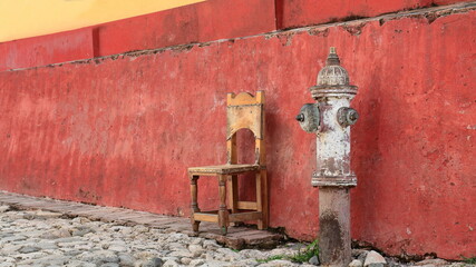 Rusty old fire hydrant and wood chair next to the red dado rail, colonial house on Calle Amargura Street, Plaza Mayor Square area. Trinidad-Cuba-268