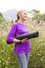 Enjoying outdoors, caucasian senior female holding a yoga mat stands in a field