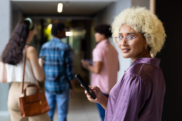 Biracial woman with glasses, blonde curls in purple top, holds smartphone in office - Powered by Adobe