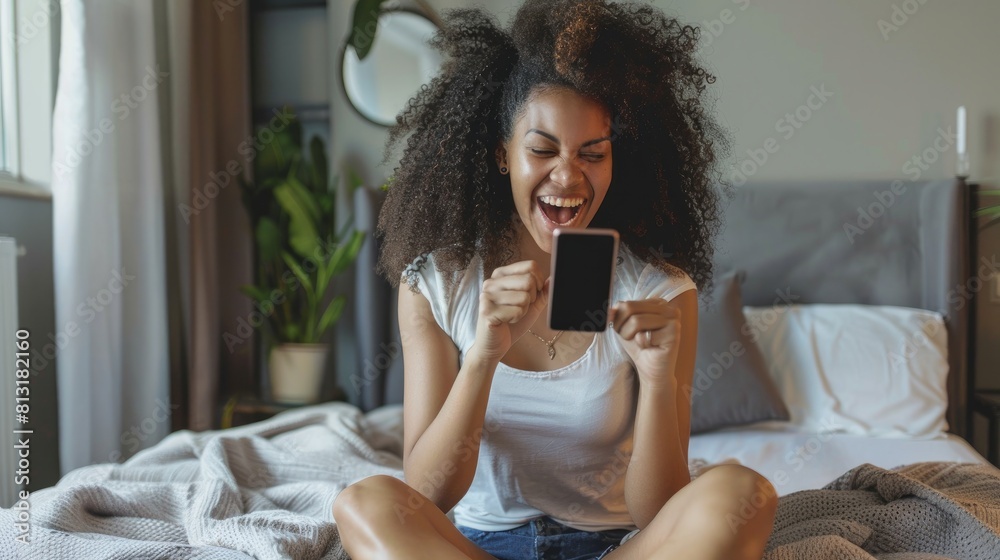 Wall mural happy young brazilian woman holding smartphone and celebrates with a clenched fist sitting on bed at