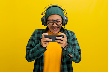Excited young Asian man, wearing headphones, a beanie hat and casual shirt, engages in playing an online mobile game on his phone while simultaneously live broadcasting the gameplay on the internet © Jamaludinyusup