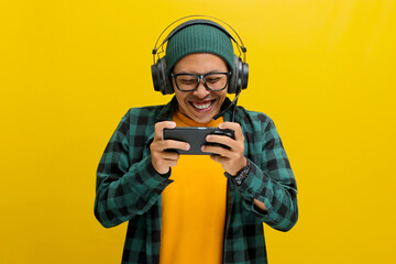 Excited young Asian man, wearing headphones, a beanie hat and casual shirt, engages in playing an...