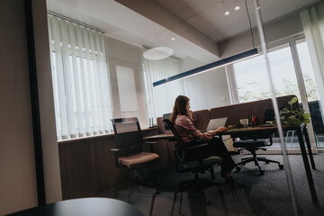 A young woman is focused on her work in a well-lit modern office room, using a laptop at her desk by large windows.