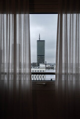 Modern glass building seen from the interior of a room through a window with curtains. It is located in the city of Warsaw, the capital of Poland