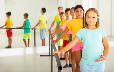 Cute cheerful preteen girl working near ballet barre during group choreography class.