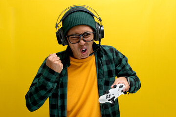 An enthusiastic Asian gamer, adorned in a beanie hat, casual shirt, and headphones, celebrates victory while playing a video game on a console with a gamepad or joystick isolated on yellow background.