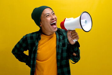 Excited Asian man in a beanie hat and casual shirt makes an announcement with a megaphone, using a loudspeaker to share news, a special offer, discount, or sale. Isolated on a yellow background.