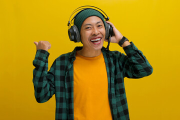 Excited Asian man in a beanie and casual clothes, wearing headphones and enjoying music or a...