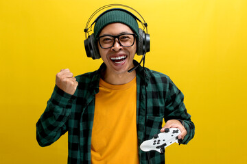 An enthusiastic Asian gamer, adorned in a beanie hat, casual shirt, and headphones, celebrates victory while playing a video game on a console with a gamepad or joystick isolated on yellow background. © Jamaludinyusup