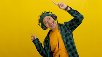 Excited Asian man in a beanie and casual clothes, wearing headphones and enjoying music or a...
