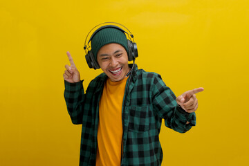Young Asian man in a beanie and casual clothes dances with joy while listening to music on headphones. Isolated on a yellow background.
