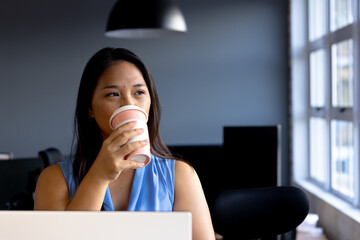At office, biracial businesswoman wearing blue, sipping coffee, looking away