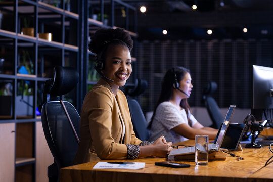 Diverse female colleagues in headsets at office, working late, focused on screens