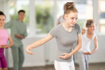 Slender teenage girl practicing ballet positions together with others in dance studio during...
