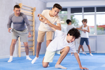 Focused teenager learning effective self defence techniques in sparring with boy under guidance of experienced instructor in gym, practicing painful armlock