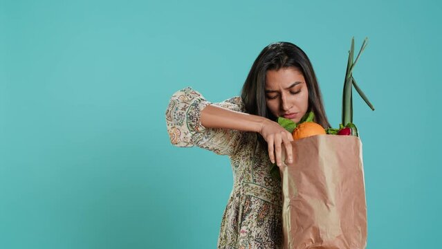 Indian woman with ecological paper bag in hands looking at bell pepper, living healthy lifestyle. Ecology lover holding shopping bag with organic produce, studio backdrop, camera A