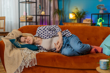 Upset young woman embracing belly suffering from stomachache lying on sofa. Unhappy sad Caucasian...