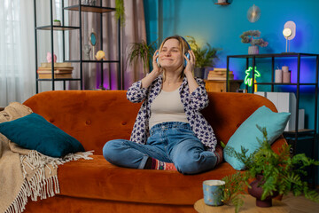 Obraz premium Happy overjoyed woman in wireless headphones relaxing sitting on couch at home apartment choosing listening favorite energetic disco dancing music. Caucasian girl enjoying weekend leisure activities.