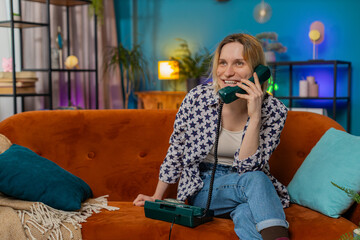 Smiling young woman making wired telephone call conversation with friends sitting on couch at home...