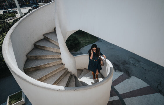 Businesswoman using smart phone while walking down a spiral outdoor stairway in a modern urban setting.