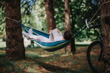 A young woman enjoys a quiet reading moment in a hammock, surrounded by trees in a serene park,...