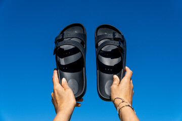 Overhead shot of a woman holding black summer sandals in the air against the blue sky.