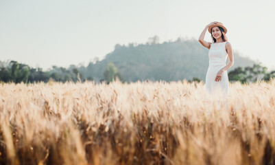 Smiling woman in a white dress and sunhat stands amidst a golden wheat field, embodying the tranquility and beauty of rural life.