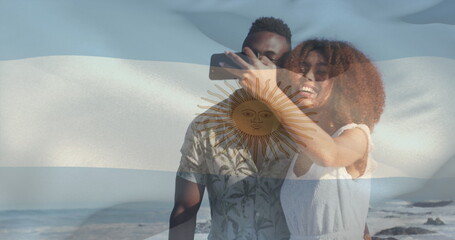 Composite image of waving argentina flag over african american couple taking a selfie at the beach