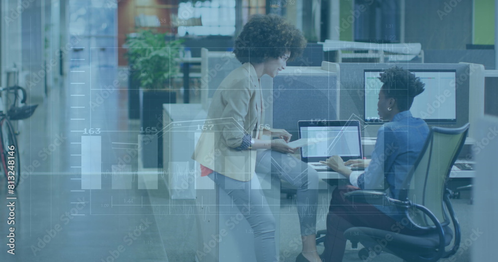 Wall mural Image of graphs and changing numbers over diverse female coworkers discussing on desk